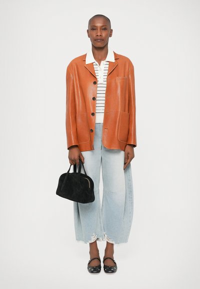 Orange Lederjacke mit Knöpfen, gestreiftes weißes Hemd, lockere hellblaue Jeans, schwarze Wildlederhandtasche und schwarze flache Schuhe mit Punkten.