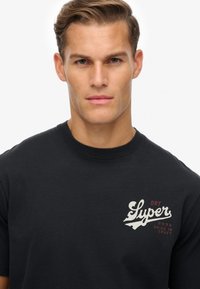 Camiseta de algodón negra con un diseño impreso en blanco y rojo en el lado izquierdo del pecho, con un cuello redondo clásico y mangas cortas.