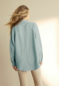 Lyseblå, tekstureret, langærmet skjorte med krave og sideslidser. Den har en afslappet pasform med detaljeret søm bagpå og afrundet kant.
