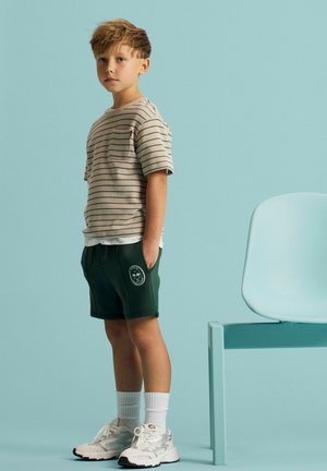 Plavokosi dječak stoji s rukama u džepovima, nosi prugastu bež košulju, tamnozelene kratke hlače, bijele čarape i bijele tenisice uz svijetloplavu stolicu.