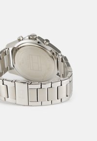 Nerūdijančio plieno laikrodis su poliruota apyranke, apvaliu korpusu ir chronografo funkcijomis; išgraviruota prekės ženklo emblema ant nugarėlės.