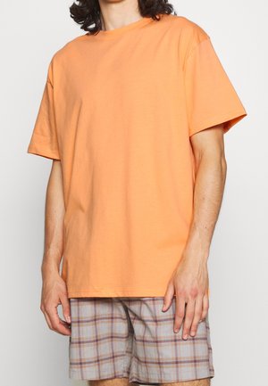Person trägt ein einfaches oranges kurzärmeliges T-Shirt und graue karierte Shorts vor neutralem Hintergrund.