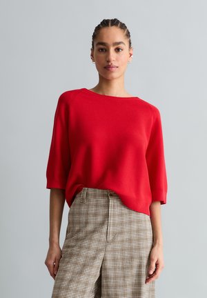 Vrouw met gevlochten haar, gekleed in een rode top met driekwart mouwen en een hoge taille geruite, bruine broek, staand tegen een eenvoudige grijze achtergrond.