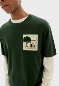 Groene katoenen t-shirt met een patchontwerp van een boom en een gebouw. Gelaagd met een witte lange mouwen shirt eronder.