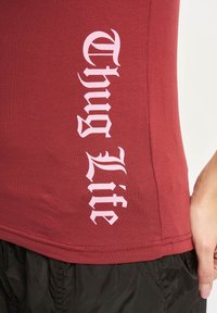 Rød tætsiddende T-shirt med lodrette pink bogstaver, der angiver "Thug Life" i en fed, stiliseret skrifttype. Glat tekstur, minimalistisk design, korte ærmer.