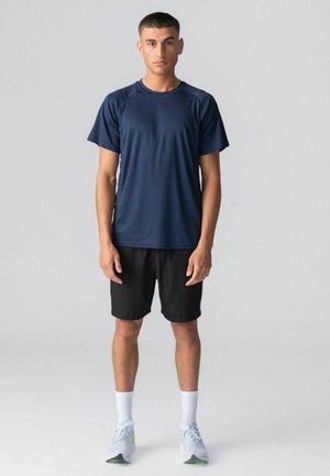 Ung mand stående iført en marineblå sportstshirt, sorte shorts, hvide sokker og hvide løbesko på en lysegrå baggrund.