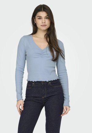 Top azzurro chiaro a maniche lunghe con scollo a V e dettaglio frontale arricciato. Lunghezza cropped con orlo ondulato e texture a coste. Abbinato a jeans scuri.