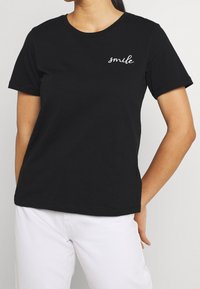 Černé bavlněné tričko s kulatým výstřihem a krátkými rukávy. Na levé straně je výšivka "Smile" v bílé kurzivě. Viditelné bílé kalhoty.