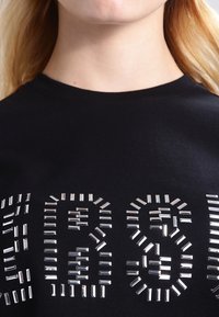  camiseta de algodón negra con letras de brillantes plateados en un diseño audaz. La camiseta tiene un cuello redondo y mangas cortas.