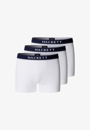 Mužský trup a nohy v bielych boxerkách Hackett s tmavomodrým pásom, stojace bosé na jednoduchom svetlosivom pozadí.