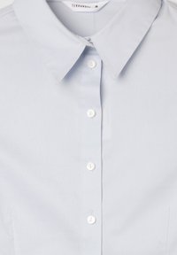 Camicia azzurra chiara con bottoni, realizzata in tessuto liscio, con colletto a punta e cinque bottoni bianchi lungo la parte anteriore. Senza fantasie o dettagli.