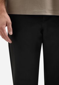 Zwarte broek met een gladde textuur en een getailleerde pasvorm. Een hand steunt tegen de stof, waardoor de details langs de zijnaad naar voren komen.