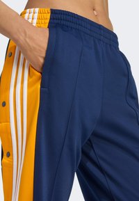 Sport pants featuring a navy blue main body, orange side panels, and three white stripes. Made from smooth, lightweight fabric with pockets.