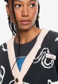 Knitted cardigan in dark blue with white designs and soft pink ribbed trim. Features a button closure and a logo patch on the left side.