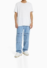 Person klädd i en enkel vit t-shirt, ljusblå vida jeans med horisontella sömmar och vita sneakers, stående mot en vit bakgrund.