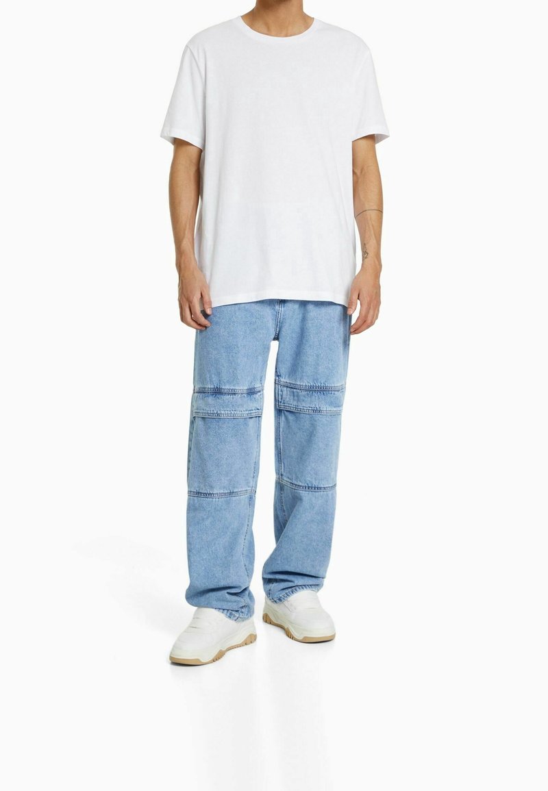 Person klädd i en enkel vit t-shirt, ljusblå vida jeans med horisontella sömmar och vita sneakers, stående mot en vit bakgrund.