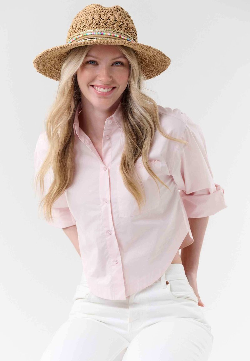 Femme souriante aux longs cheveux blonds portant un chapeau de paille, une chemise rose clair boutonnée et un pantalon blanc sur un fond uni.