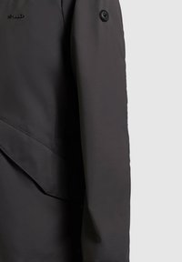 Must detailne pikem varrukaga jakk, näidates varrukat, klapiga taskut, ümmargust musta tähnaga nuppu ja väikest musta tikitud brändi logo.