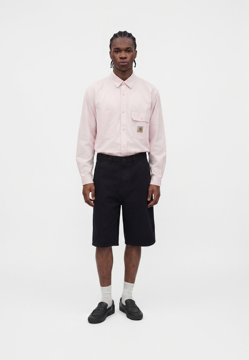 Männliches Model trägt ein hellrosa langärmeliges Hemd mit Brusttaschen, schwarze knielange Shorts, weiße Socken und schwarze Loafers, steht aufrecht.