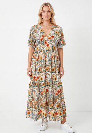 Blonde woman wearing a floral V-neck maxi dress with puff sleeves and white sneakers, standing against a plain white background.