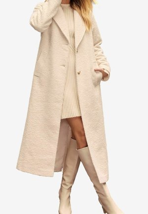 Woman wearing long beige wool coat, ribbed knit dress, and knee-high beige boots, standing with one hand in pocket and one touching hair.