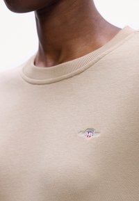 Gros plan d'une personne portant un t-shirt beige à col rond avec un petit logo brodé représentant un bouclier avec des étoiles et des rayures sur la poitrine.