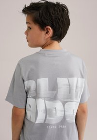 Pelēka T-krekls ar lielu baltu grafisku tekstu uz muguras, kurā rakstīts "BLU RID" un zem tā "SINCE 1986". Mīksts kokvilnas materiāls.
