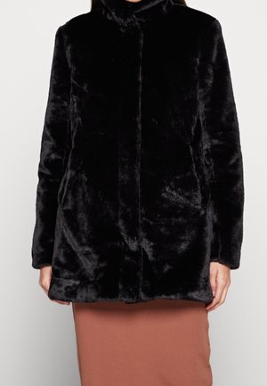 Frau trägt einen schwarzen Kunstpelzmantel mit hohem Kragen, kombiniert mit einem braunen, gerippten Rock vor einem einfarbigen Hintergrund.