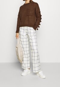 Pull en tricot marron avec une poche et des accents circulaires aux manches, associé à un pantalon large à carreaux blancs et des baskets blanches. Portant un sac beige.