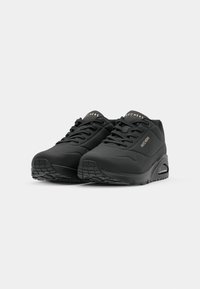 Paire de baskets noires Skechers à lacets avec semelles épaisses et amorti à air visible sur un fond clair uni.
