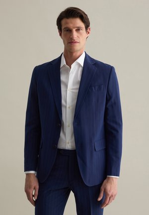 Navy pinstripe suit with a two-button closure, notch lapels, and a white dress shirt. Tailored fit with side pockets. Smooth fabric texture.