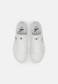 Bílé plátěné tenisky s šněrovacím designem, s černými logovými akcenty po stranách a texturovanou bílou gumovou podrážkou.