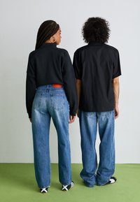 To personer står ryggen til ryggen, iført en sort cropped sweatshirt og en kortermet sort skjorte, kombinert med høyt liv blå jeans.