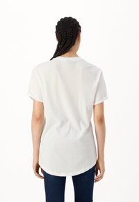 Biała koszulka bawełniana z podwiniętymi krótkimi rękawami, okrągłym dekoltem i nieco dłuższym tyłem, noszona z ciemnoniebieskimi dżinsami.