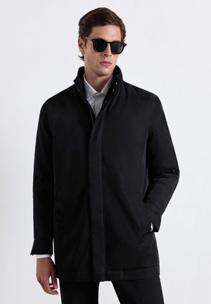 Junger Mann trägt schwarze Jacke, weißes Hemd und schwarze Sonnenbrille, mit einer Hand in der Jackentasche, steht vor einem einfarbigen Hintergrund.