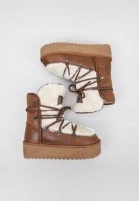 Botas de piel sintética marrón con forro de piel sintética color crema, suelas de goma marrón gruesas y cordones decorativos marrones. Presentan una puntera redonda y un cuello acolchado.