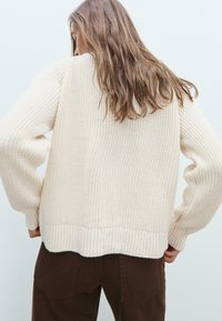 Pull en tricot couleur crème avec des poignets et un ourlet côtelés, affichant une coupe décontractée et un tissu texturé. Porté sur un pantalon marron, vu de dos.