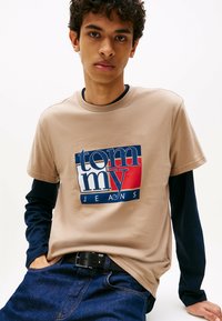Ung mand iført lagdelte beige og marineblå skjorter med "Tommy Jeans" logo, iført mørkeblå jeans og sort bælte, stående mod en hvid baggrund.