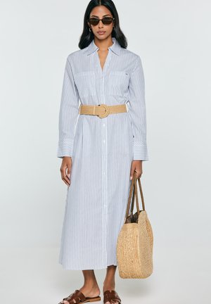 Kobieta w długiej białej i niebieskiej sukience koszulowej w paski, z beżowym plecionym paskiem, brązowych sandałach, okularach przeciwsłonecznych, trzymająca plecioną torbę.
