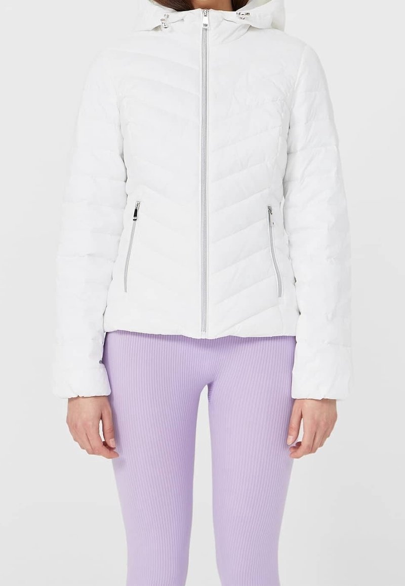 Femme vêtue d'une veste matelassée blanche avec fermeture éclair devant et poches latérales zippées, associée à un legging côtelé violet clair.