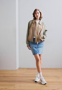 Giubbotto bomber leggero in beige e verde oliva, abbinato a una gonna di jeans. Indossato con sneakers bianche e calzini alla caviglia. Texture liscia.