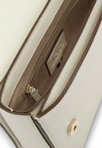 Interno della borsa in pelle di colore crema con dettagli marroni. Presenta una tasca con zip, etichetta di marca e una chiusura a scatto. Superficie testurizzata.