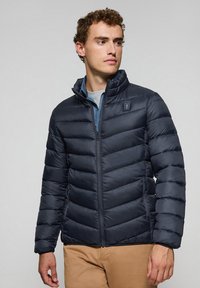 Chaqueta acolchada aislante de color azul oscuro con diseño de acolchado horizontal, cuello alto, cremallera frontal y parche de logo en la parte superior izquierda del pecho.