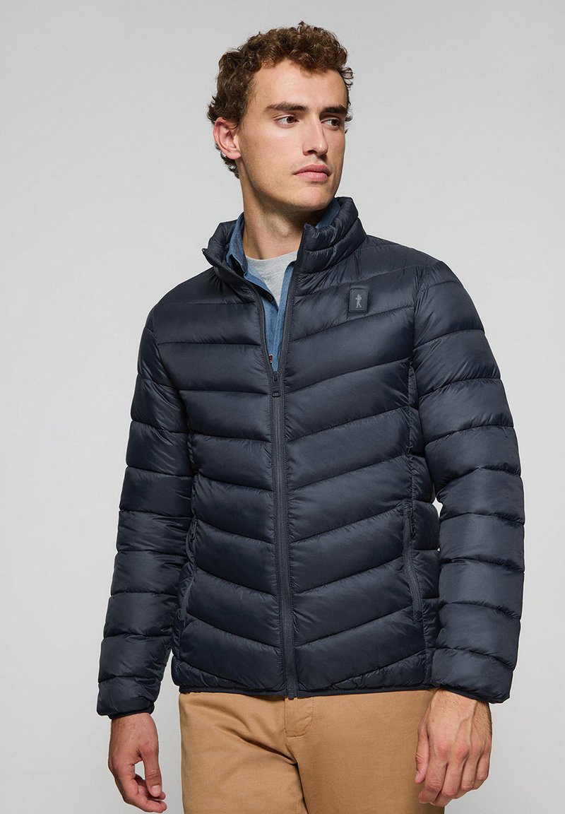 Chaqueta acolchada aislante de color azul oscuro con diseño de acolchado horizontal, cuello alto, cremallera frontal y parche de logo en la parte superior izquierda del pecho.
