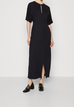 Woman wearing a long black dress with short sleeves, a front slit, and black heeled loafers with metal chain detail.