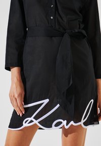 Vestido negro con mangas largas, cierre de botones, cinturón a la cintura y bordado blanco de "Karl" en el dobladillo. Textura de tela suave.