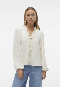 Blouse couleur crème à manches longues, avec un col froncé, en tissu texturé, et une coupe décontractée, assortie à un jean en denim bleu.