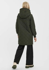 Parka verde trapuntata con cappuccio, caratterizzata da un taglio lungo, spalle scese e cerniere laterali. Indossata sopra pantaloni neri a gamba larga.
