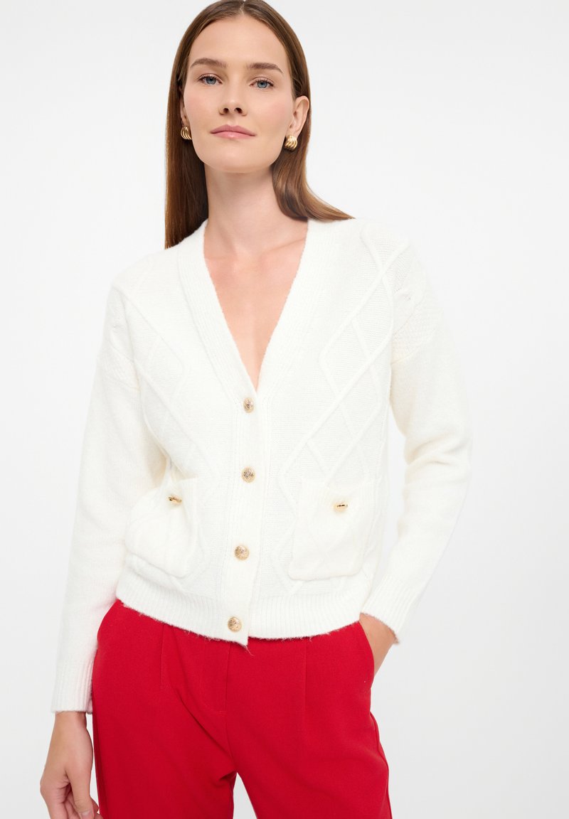 Witte gebreide cardigan met een diepe V-hals, een ruitpatroon, voorzakken en gouden knopen. Gecombineerd met rode broeken.
