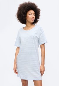 Γυναίκα με κατσαρά μαλλιά που φοράει ένα φαρδύ, κοντομάνικο φόρεμα με γαλάζιες και λευκές ρίγες, κοιτάζοντας ελαφρώς προς το πλάι.